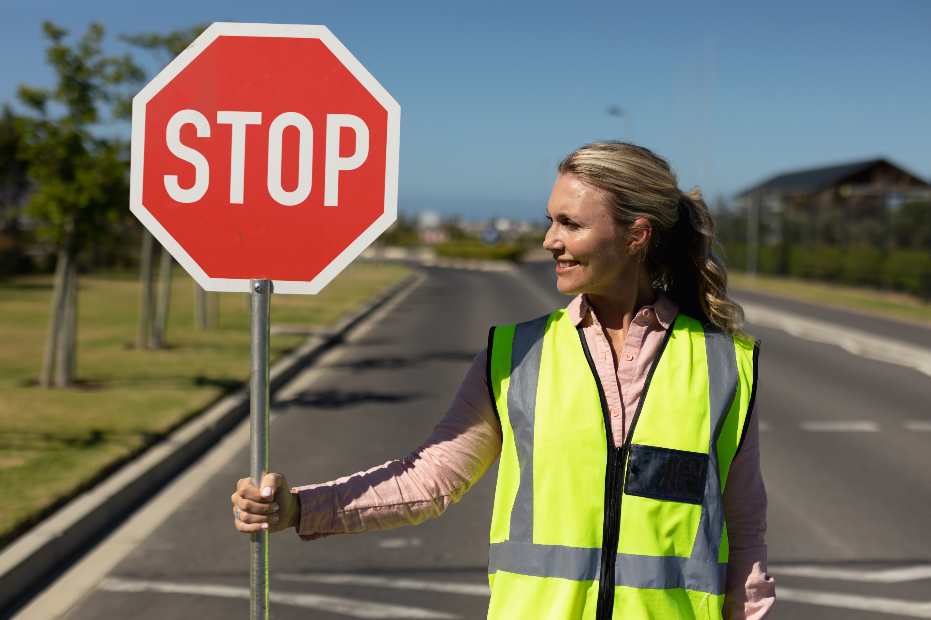 Person mit langen blonden Haaren und gelber Warnweste steht auf einer Straße und hält ein Stoppschild in der Hand