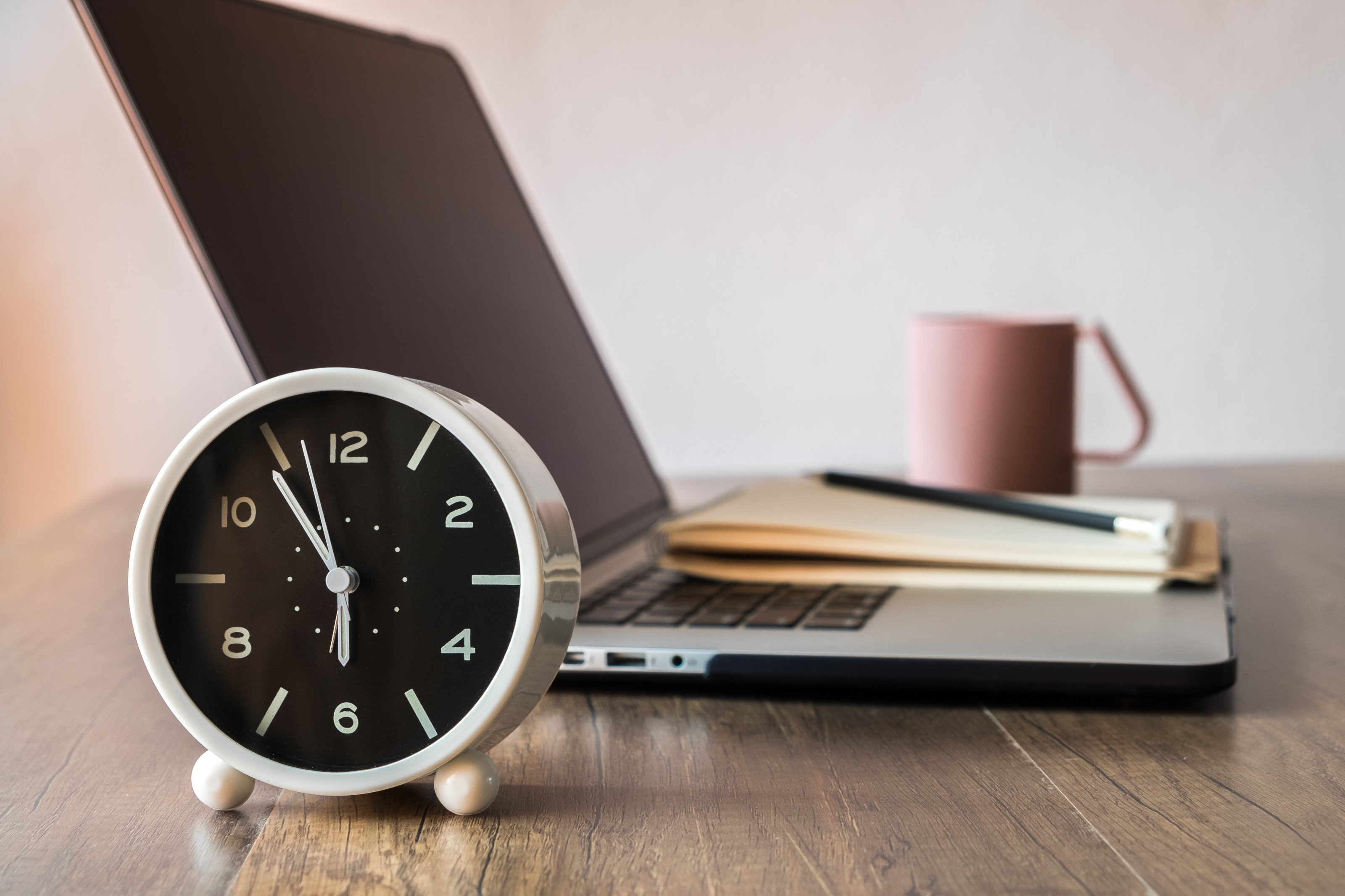 Wecker mit Uhrzeit kurz vor 06:00 Uhr steht vor einem aufgeklapptem Laptop mit Notizblock, Stift und Tasse auf einem Holztisch