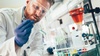 Person mit kurzen Haaren und Bart trägt eine weiße Schutzkleidung mit Brille und blauen Handschuhen und blickt auf ein Reagenzglas mit Flüssigkeit in der Hand während im Hintergrund weitere Reagenzgläser mit Flüssigkeiten in einem Labor stehen