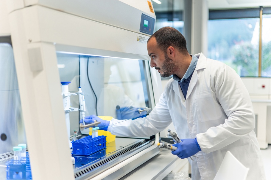 Seitliche Aufnahme einer Person mit weißem Kittel, Bart, dunklen Haaren und Handschuhen. Die Person steht an einer Station mit einer Glasscheibe, hinter der Reagenzgläser stehen