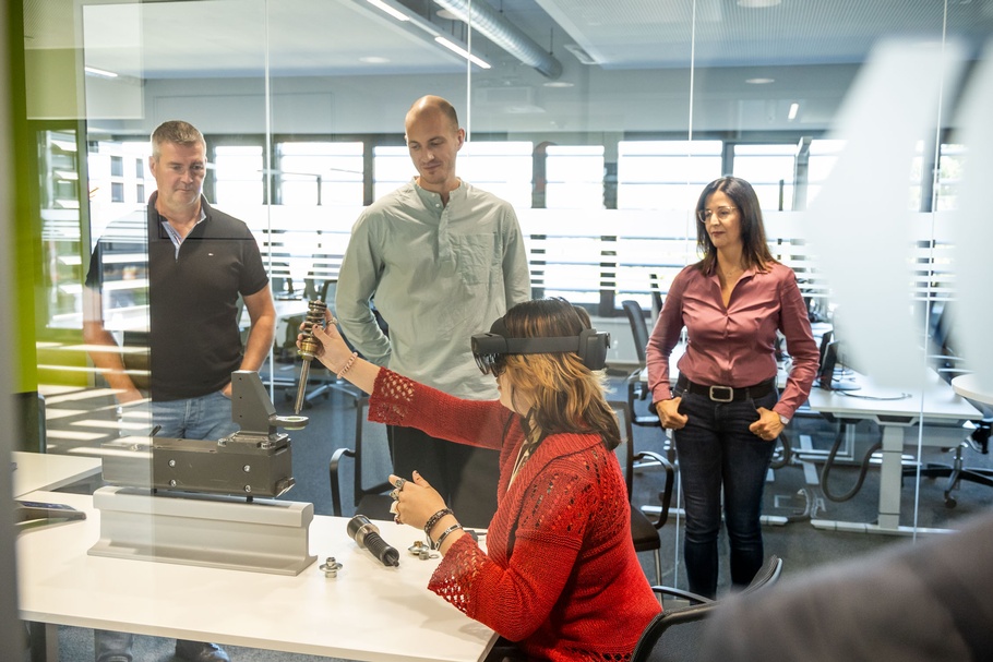 Personen im Büro. Eine davon trägt eine VR-Brille, die anderen schauen dabei zu.
