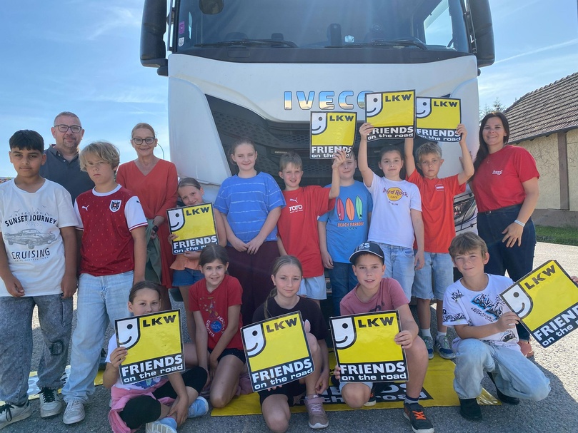 Schulklasse mit Erwachsenen vor einem weißen LKW; Kinder präsentieren Schilder mit „LKW – Friends on the road“.