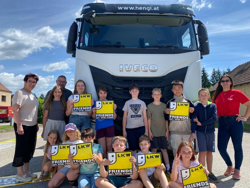 Schulklasse mit Erwachsenen vor einem weißen LKW; Kinder präsentieren Schilder mit „LKW – Friends on the road“.