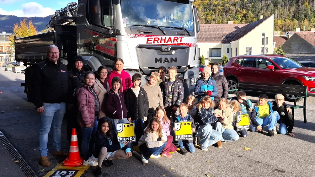 Gruppe von Kindern einer Polizistin und erwachsenen Personen steht vor einem großen LKW mit der Aufschrift ‚Erhart‘. Einige Kinder halten gelbe Schilder mit der Aufschrift ‚LKW Friends on the Road‘.