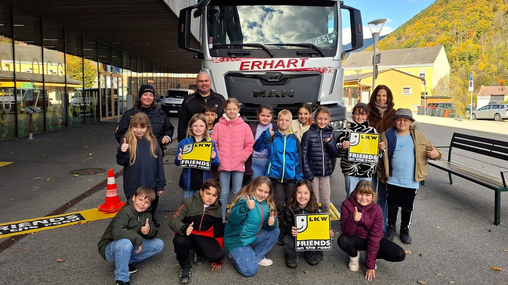 Gruppe von Kindern einer Polizistin und erwachsenen Personen steht vor einem großen LKW mit der Aufschrift ‚Erhart‘. Einige Kinder halten gelbe Schilder mit der Aufschrift ‚LKW Friends on the Road‘.