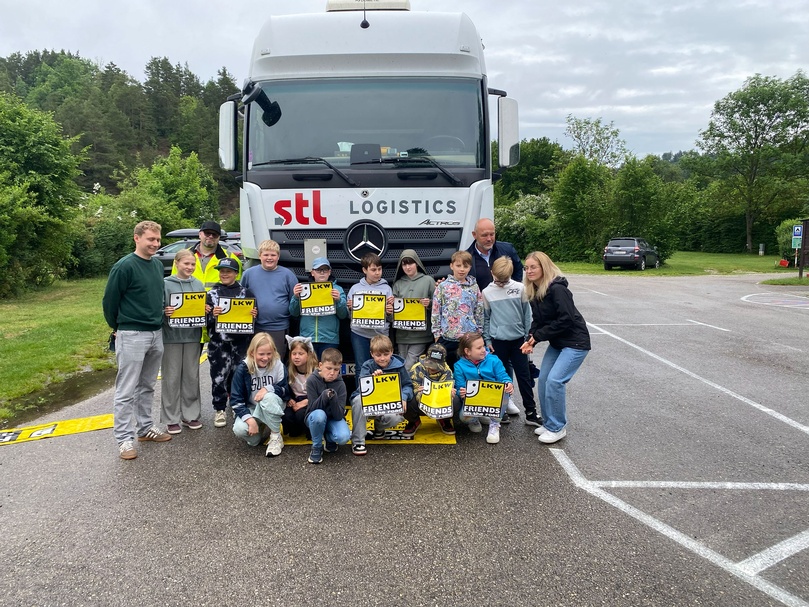 Gruppe von Schulkindern und Begleitpersonen steht vor einem weißen Lkw mit „stl logistics“-Logo; mehrere Kinder halten gelbe Warnmaterialien mit der Aufschrift „LKW FRIENDS“; Aufnahme im Freien auf einem Parkplatz.