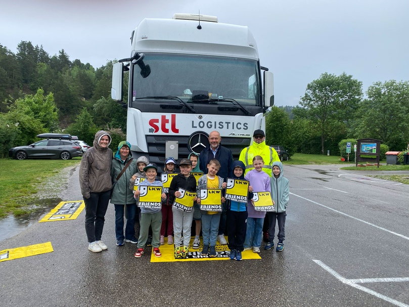 Gruppe von Schulkindern und Begleitpersonen steht vor einem weißen Lkw mit „stl logistics“-Logo; mehrere Kinder halten gelbe Warnmaterialien mit der Aufschrift „LKW FRIENDS“; Aufnahme im Freien auf einem Parkplatz.