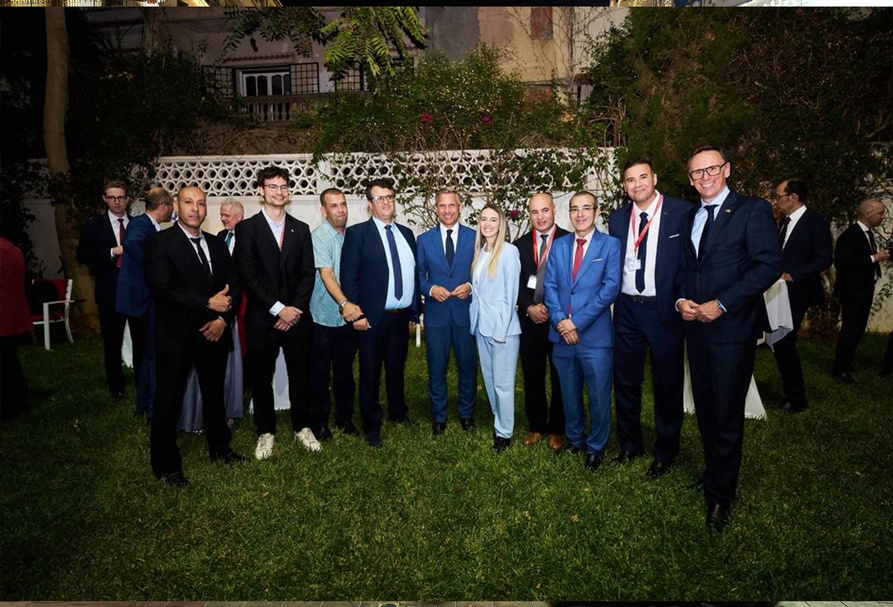 Porträt von neun Personen, die nebeneinander auf einer Rasenfläche stehen. Hinter der Gruppe befinden sich eine weiße Mauer mit geometrischen Öffnungen, Sträucher und Bäume. Im Hintergrund stehen weitere Personen neben weiß gedeckten Stehtischen