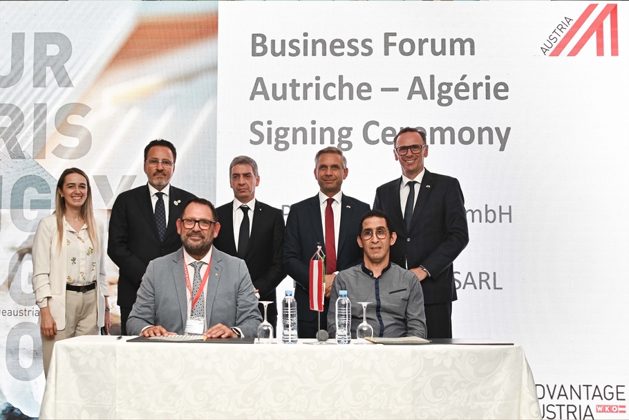 An einem Tisch sitzen zwei Personen. Vor beiden Personen liegt jeweils eine aufgeschlagene Mappe. Hinter ihnen stehen fünf weitere Personen, hinter denen eine Leinwand ist, auf der unter anderem Businessforum Österreich Algerien steht