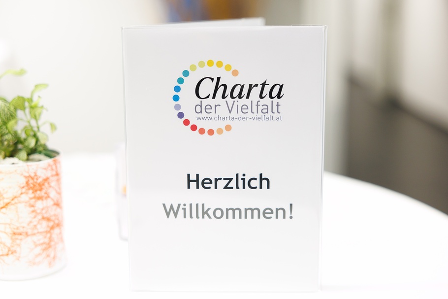 Das Foto zeigt einen weißen, rechteckigen Willkommensaufsteller in einer transparenten Halterung auf einem Tisch mit weißem Tischtuch. Im oberen Bereich befindet sich das Logo „Charta der Vielfalt“. Der Schriftzug „Charta“ ist in schwarzer, geschwungener Schrift gestaltet