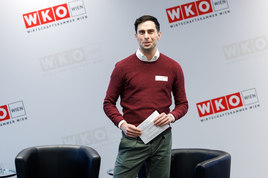 Eine Person mit kurzen, dunklen Haaren steht in einem Innenraum vor einer hellgrauen Wand mit mehreren Logos der Wirtschaftskammer Wien. Sie trägt weißes Namensschild auf der Brust und hält mehrere Zettel in den Händen