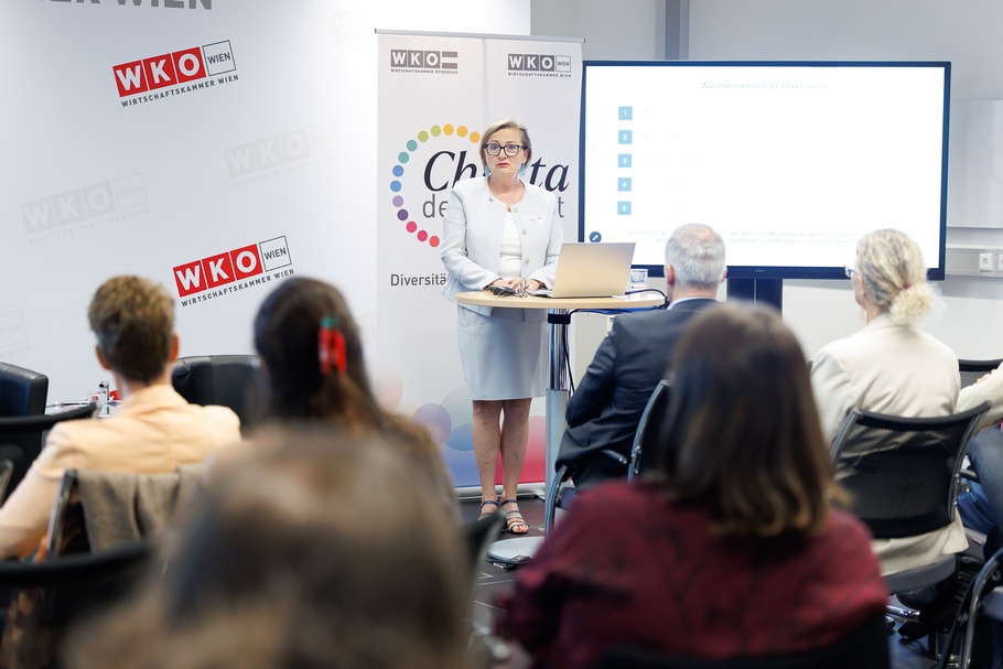 Eine Person mit kurzen, blonden Haaren und Brille steht vor mehreren sitzenden Personen in einem Veranstaltungsraum. Sie trägt ein Namensschild