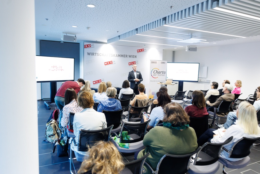 Heller Konferenzraum mit mehreren Reihen schwarzer Stühle, auf denen zahlreiche Personen sitzen. Die meisten sind von hinten abgebildet und blicken nach vorne. Im vorderen Bereich des Raums steht eine Person mit kurzen, grauen Haaren. Hinter der vortragenden Person befindet sich eine helle Wand mit mehreren rot-weißen Logos der Wirtschaftskammer Wien und dem Schriftzug „WIRTSCHAFTSKAMMER WIEN“