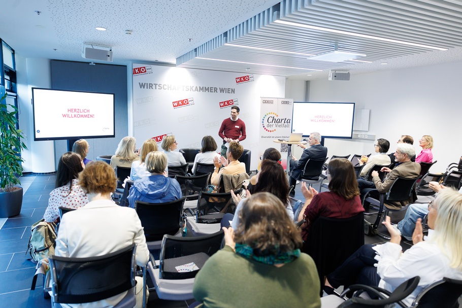 Heller Konferenzraum mit mehreren Reihen schwarzer Stühle, auf denen zahlreiche Personen sitzen. Die meisten sind von hinten abgebildet und blicken nach vorne. Mehrere Personen haben die Hände zum Applaudieren erhoben.<br />
Im vorderen Bereich des Raums steht eine Person mit kurzen, dunklen Haaren und Namensschild
