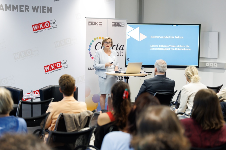 Eine Person mit kurzen, blonden Haaren und Brille steht vor mehreren sitzenden Personen in einem Veranstaltungsraum. Sie trägt ein Namensschild. Auf einem Bildschirm neben ihr steht Kulturwandel im Fokus