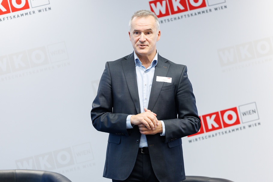 Person mit kurzen, grauen Haaren steht vor einer hellgrauen Wand mit mehreren Logos der Wirtschaftskammer Wien.<br />
Sie trägt einen dunkelgrauen Anzug mit einem hellblauen Hemd und ein weißes Namensschild am Sakko