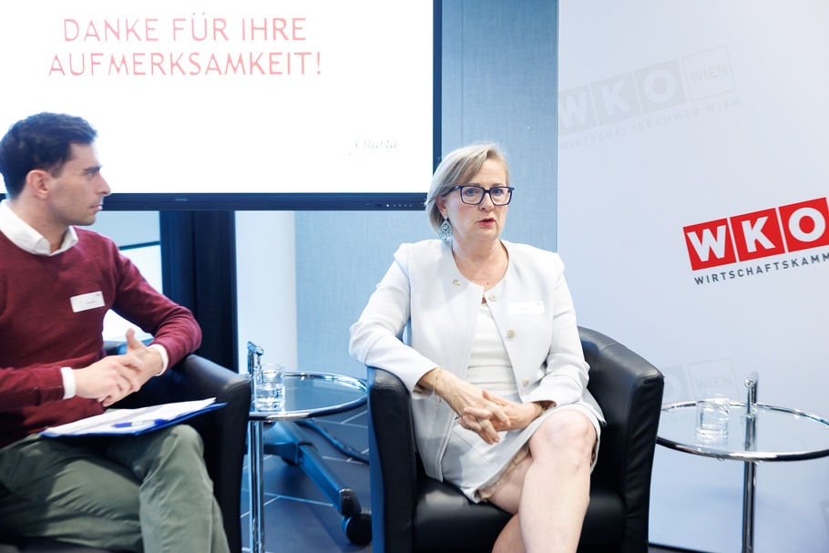 Zwei Personen sitzen in schwarzen Ledersesseln nebeneinander in einem Veranstaltungsraum. Zwischen den Sesseln und rechts daneben stehen kleine, runde Glastische mit Wassergläsern und Wasserflaschen. Im Hintergrund befindet sich ein großer Bildschirm mit der Aufschrift „DANKE FÜR IHRE AUFMERKSAMKEIT!“ in hellroten Großbuchstaben. Rechts steht eine helle Wand mit mehreren Logos und der Aufschrift „WKO Wien – Wirtschaftskammer Wien“