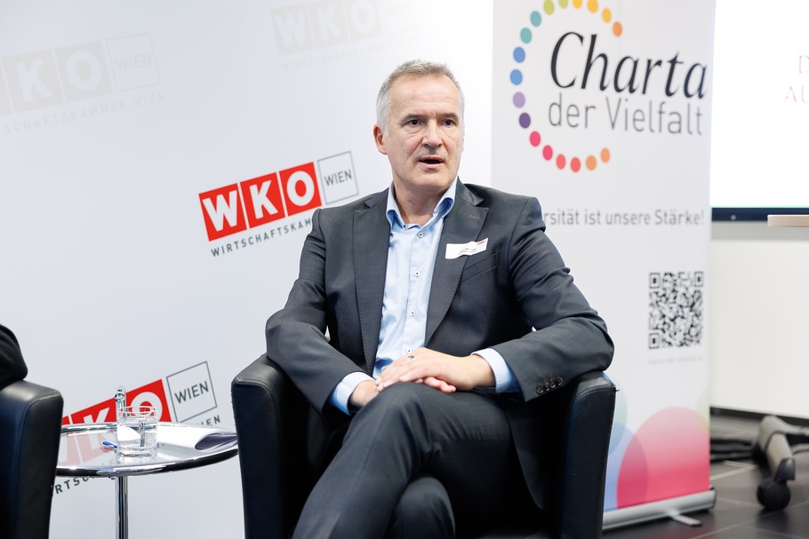 Eine Person mit kurzen, grauen Haaren sitzt in einem schwarzen Ledersessel in einem Veranstaltungsraum. Im Hintergrund befindet sich eine helle Wand mit mehreren rot-weißen Logos und der Aufschrift „WKO Wien – Wirtschaftskammer Wien“