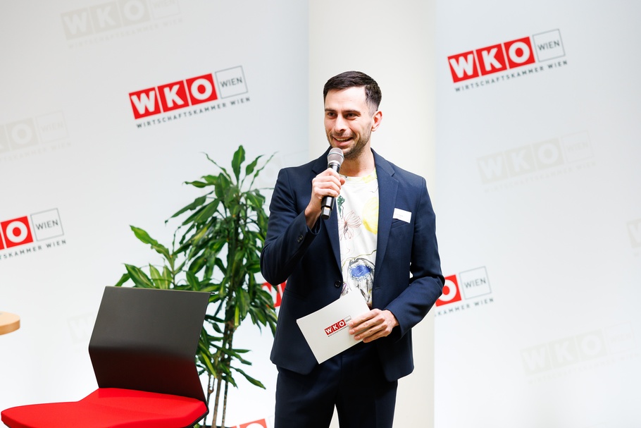 Stehende Person spricht in Mikrofon vor großer Wand mit rotweißem Logo WKO und Schriftzug Wirtschaftskammer Wien, hält in einer Hand Kärtchen mit rotweißem Logo WKO, daneben rotschwarzer Sessel und Zimmerpflanze
