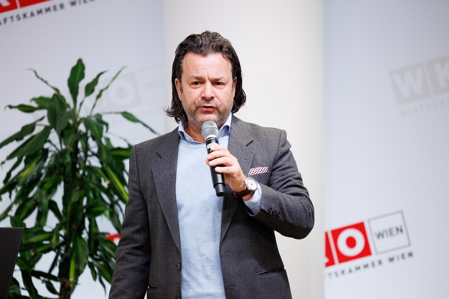 Stehende Person spricht in Mikrofon vor großer Wand mit rotweißen Logos WKO und Schriftzügen Wirtschaftskammer Wien, im Hintergrund verschwommen Zimmerpflanze