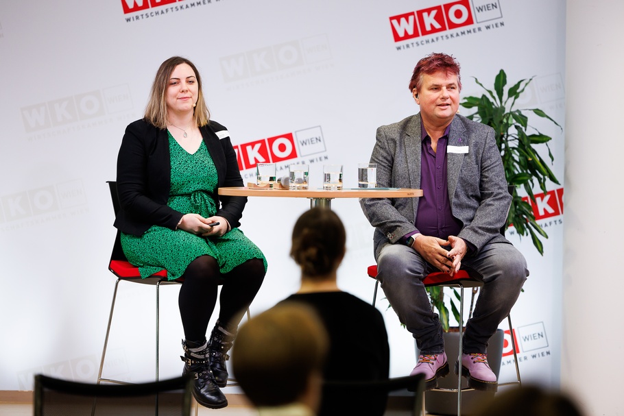 Zwei Personen mit Headmikrofonen sitzen auf erhöhten Stühlen neben Stehtisch auf Bühne vor Wand mit rotweißen Logos WKO und Schriftzügen Wirtschaftskammer Wien, im Vordergrund verschwommen weitere Person in Rückenansicht
