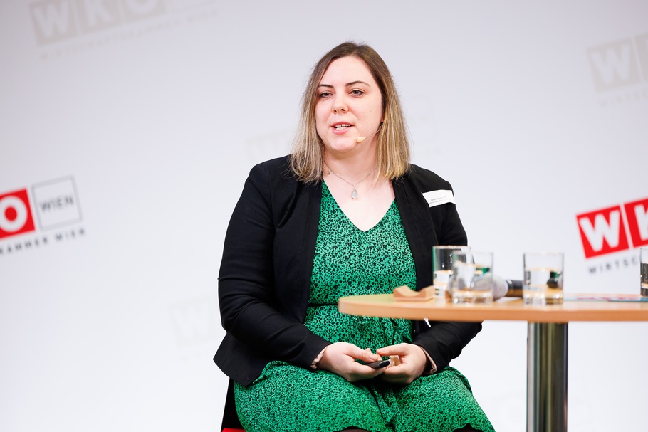 Person mit Headmikrofon sitzt auf erhöhtem Stuhl neben Stehtisch auf Bühne vor Wand mit rotweißen Logos WKO und Schriftzügen Wirtschaftskammer Wien