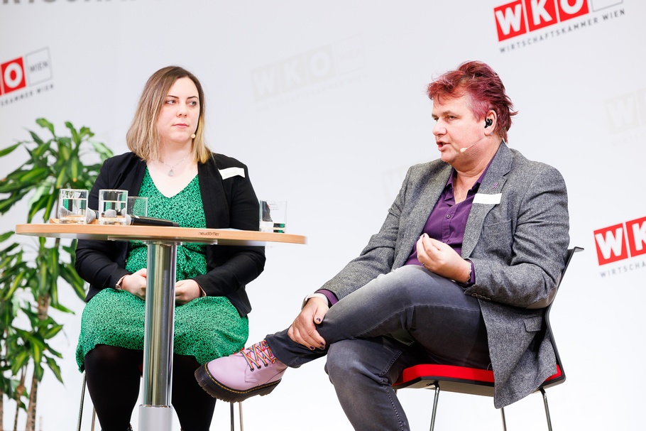 Zwei Personen mit Headmikrofonen sitzen auf erhöhten Stühlen neben Stehtisch auf Bühne vor Wand mit rotweißen Logos WKO und Schriftzügen Wirtschaftskammer Wien, eine der Personen spricht