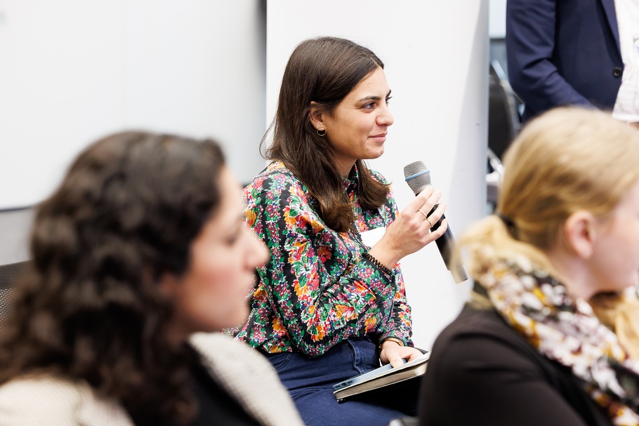 Sitzende Person im Fokus, die Mikrofon zum Mund hält, im Vordergrund verschwommen Köpfe weiterer Personen