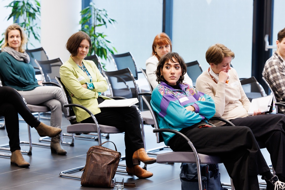 Mehrere Personen in Reihen auf schwarzen Stühlen sitzend zur Seite blickend, einige Stühle leer