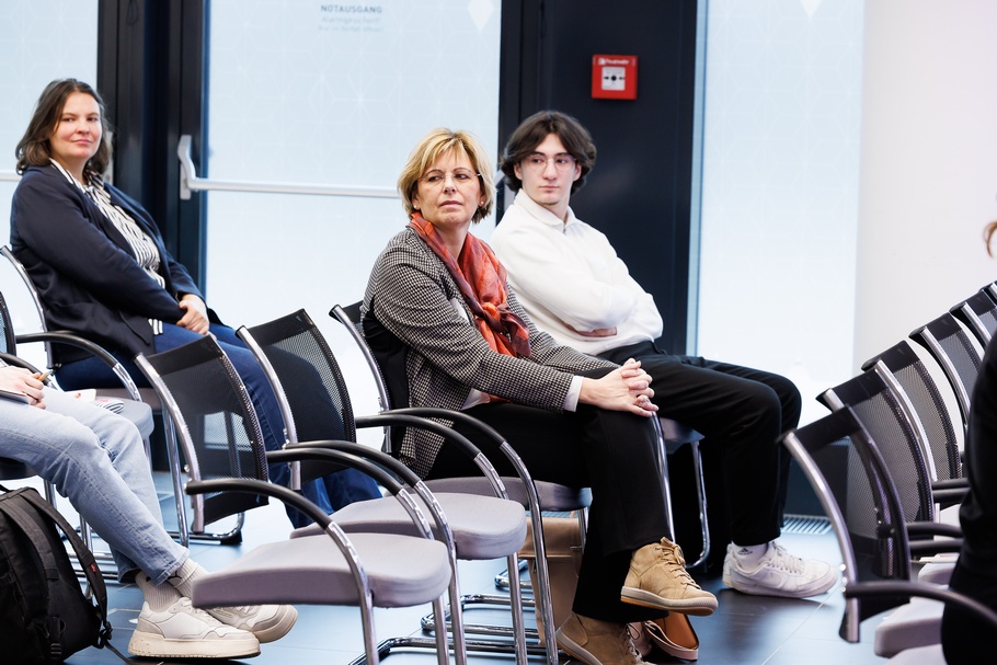 Mehrere Personen in Reihen auf schwarzen Stühlen sitzend zur Seite blickend, einige Stühle leer