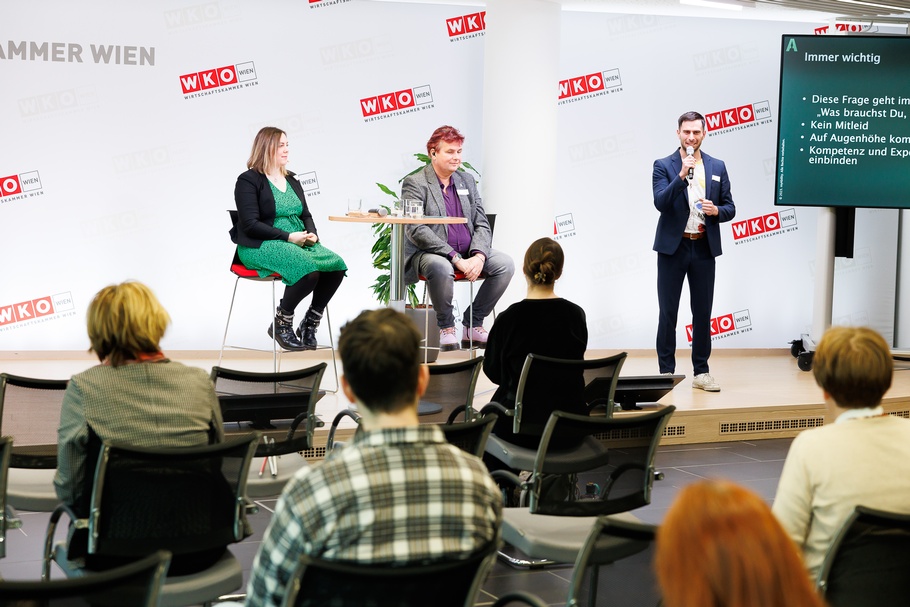 Zwei Personen mit Headmikrofonen sitzen auf erhöhten Stühlen neben Stehtisch, eine Person nebenstehend spricht in Mikrofon, auf Bühne vor Wand mit rotweißen Logos WKO und Schriftzügen Wirtschaftskammer Wien, im Vordergrund verschwommen weitere Personen sizend in Rückenansicht