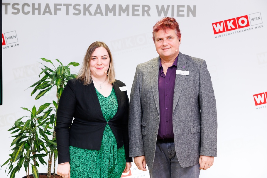 Zwei Personen mit Headmikrofonen nebeneinander auf Bühne stehend vor Wand mit rotweißen Logos WKO und Schriftzügen Wirtschaftskammer Wien, im Hintergrund verschwommen Zimmerpflanze