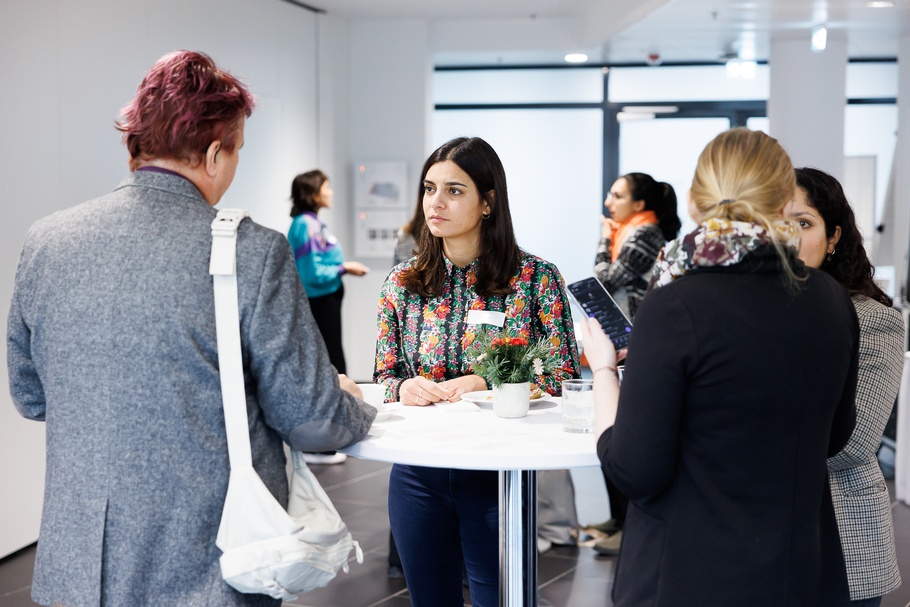Mehrere Personen um Stehtisch versammelt in Rück- und Seitenansichten, in Gesprächen miteinander, im Hintergrund weitere Personen