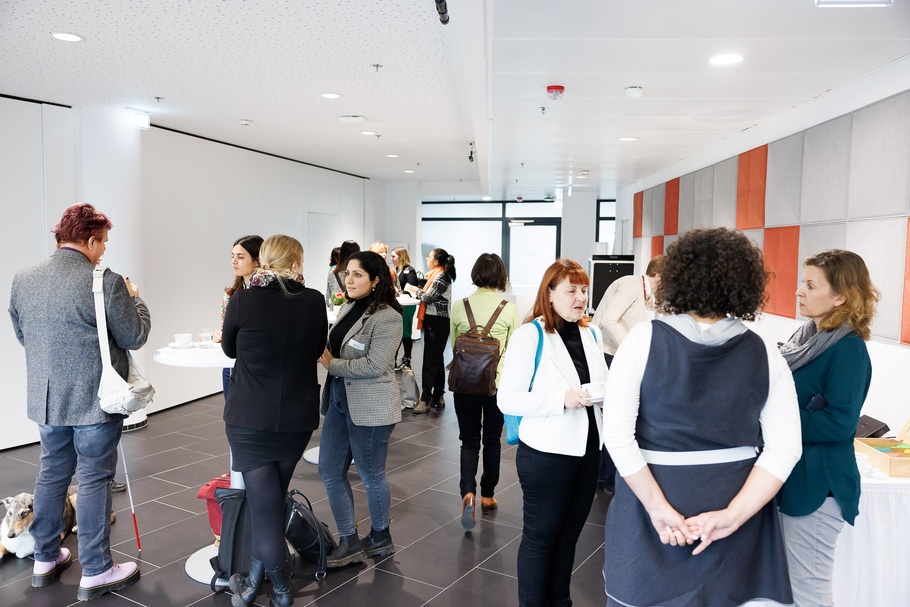 Mehrere Personen um Stehtische versammelt in Rück- und Seitenansichten, in Gesprächen miteinander, im Hintergrund Wand mit orangen und grauen Quadraten und weiße Wand sowie Glastüre