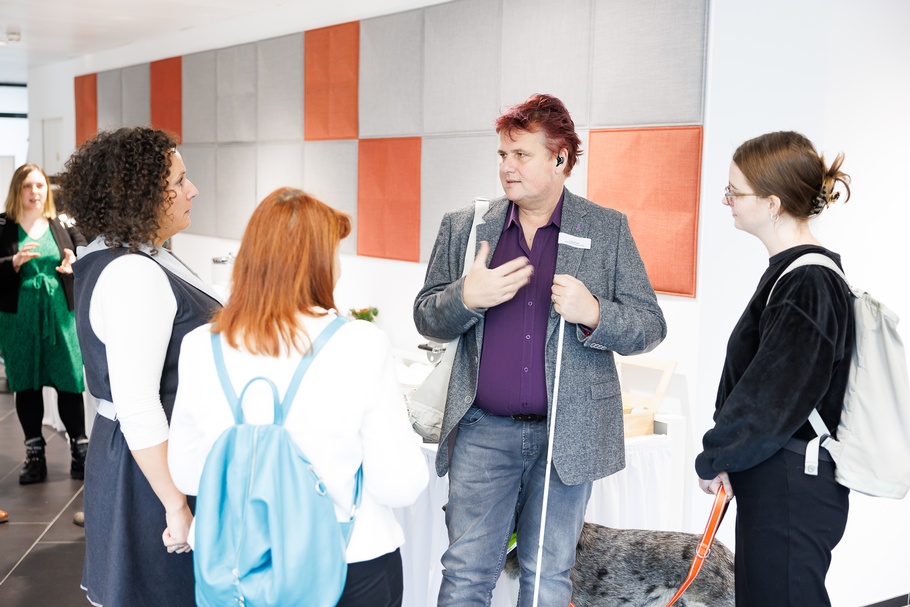 Mehrere Personen Rück- und Seitenansichten, im Gespräch miteinander, im Hintergrund Wand mit orangen und grauen Quadraten