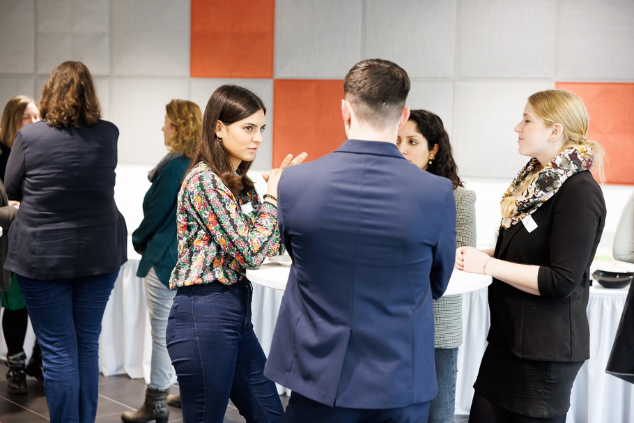 Mehrere Personen um Stehtische versammelt in Rück- und Seitenansichten, in Gesprächen miteinander, im Hintergrund Wand mit orangen und grauen Quadraten