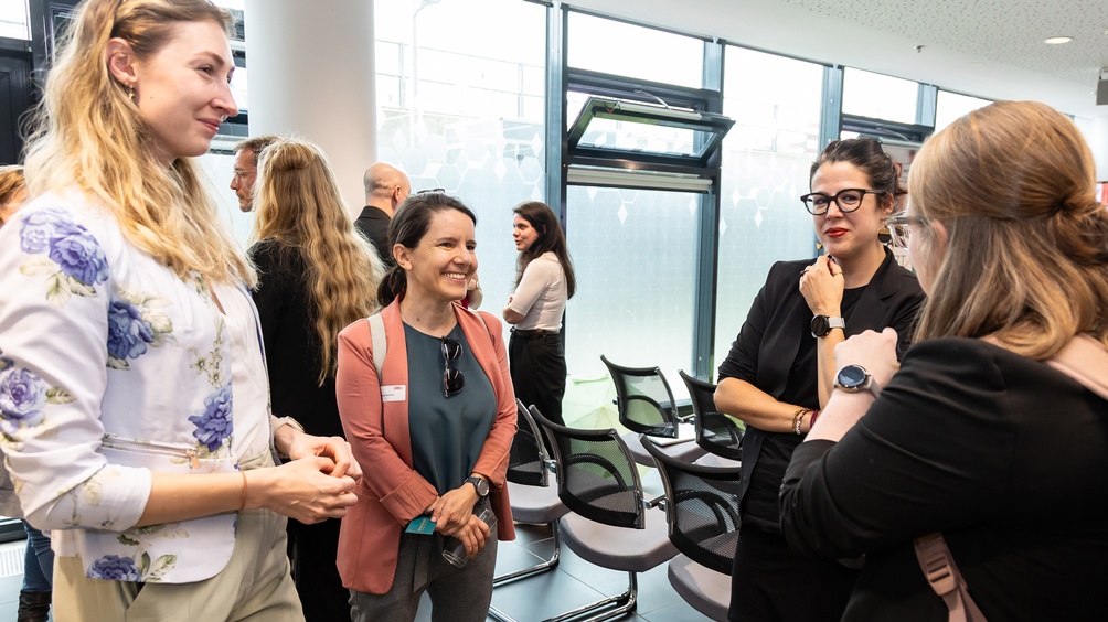 Vier Personen im Gespräch miteinander, im Hintergrund weitere Personen und leere Stühle