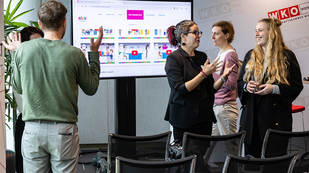 Mehrere Personen in Zweiergruppen im Gespräch in einem Büroraum stehend, im Hintergrund Bildschirm, im Vordergrund leere Stühle