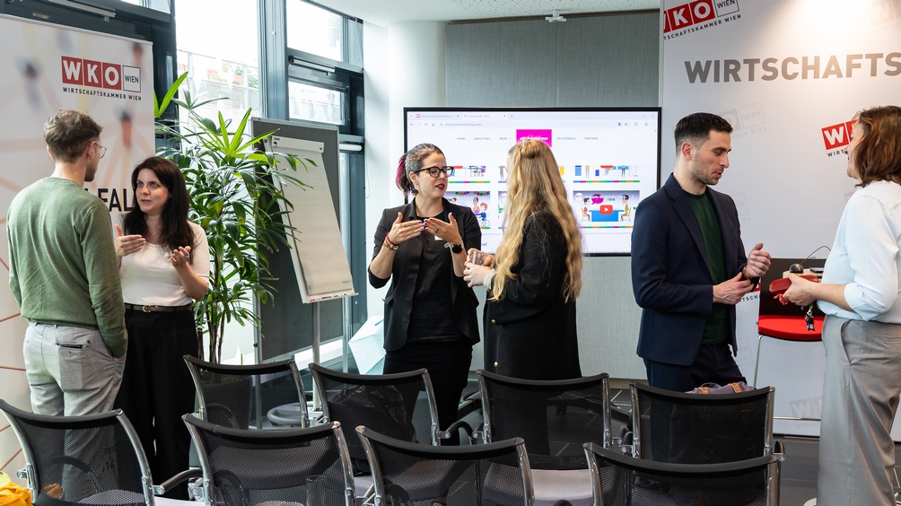 Mehrere Personen in Zweiergruppen im Gespräch in einem Büroraum zwischen Stühlen stehend, auf Rollup und Wand Logo der Wirtschaftskammer Wien