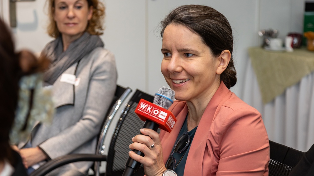 Porträt einer Person in leicht seitlicher Ansicht, die in ein Mikrofon spricht, im Hintergrund verschwommen weitere Person