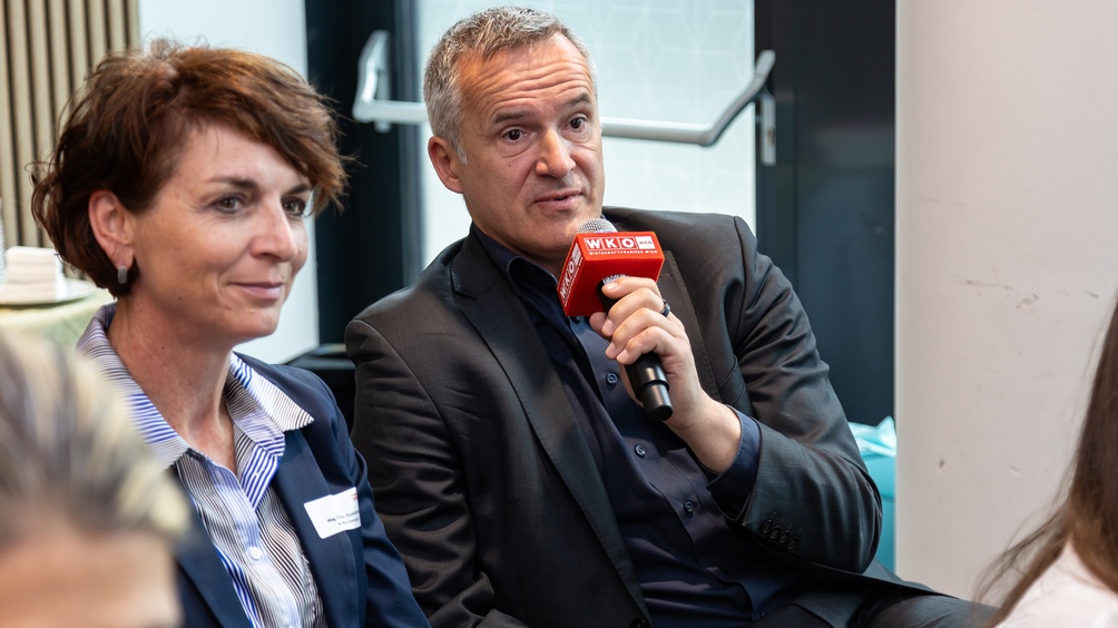 Porträt einer Person, die in ein Mikrofon spricht, daneben weitere Person