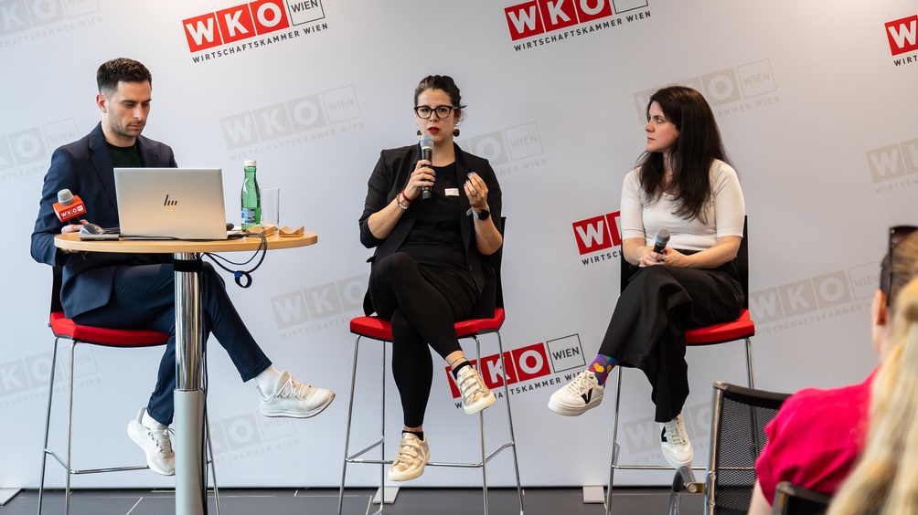 Drei Personen sitzen auf hohen Stühlen auf einer Bühne, die Person links blickt in einen aufgeklappten Laptop, die Person in der Mitte spricht in ein Mikrofon, im Hintergrund an der Wand sich wiederholendes Logo der Wirtschaftskammer Wien