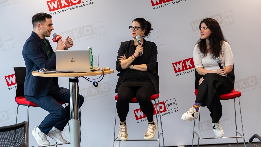 Drei Personen sitzen auf hohen Stühlen auf einer Bühne, die Person links sitzt vor einem aufgeklappten Laptop und spricht in ein Mikrofon, im Hintergrund an der Wand sich wiederholendes Logo der Wirtschaftskammer Wien