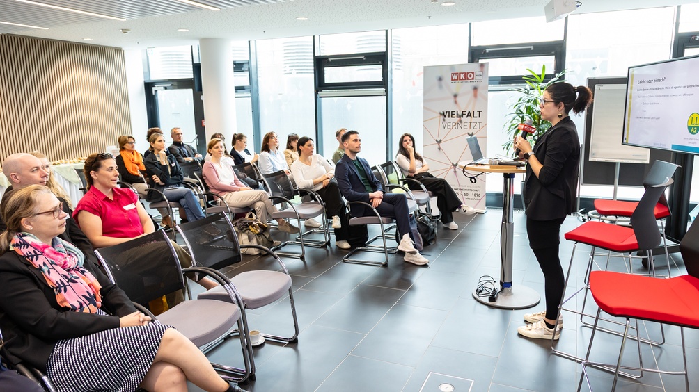 Person in Seitenansicht steht auf einer Bühne und spricht in ein Mikrofon einem sitzenden Publikum zugewandt, im Hintergrund Rollup mit Schriftzug Vielfalt vernetzt
