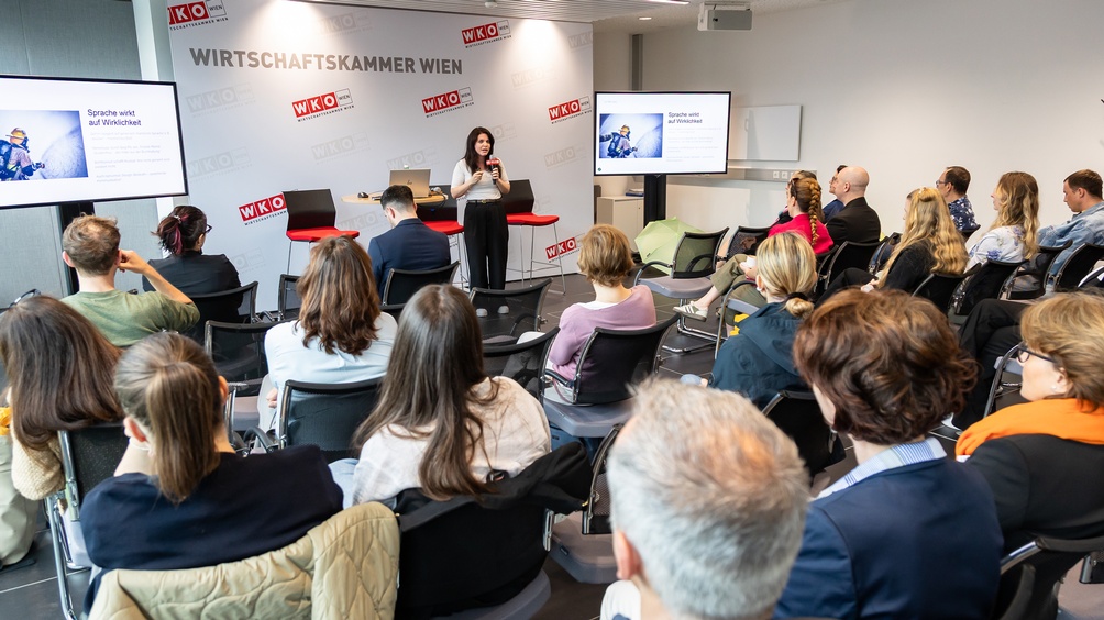  Person steht auf einer Bühne und spricht in ein Mikrofon einem sitzenden Publikum zugewandt, auf Bildschirm Schriftzug Sprache wirkt auf Wirklichkeit