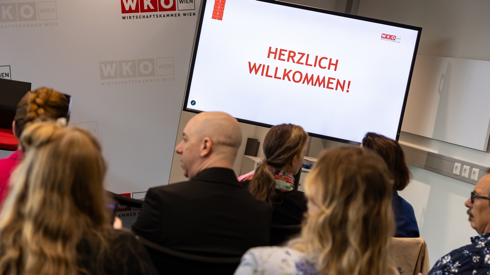 Mehrere Personen in Rückenansicht auf einer Veranstaltung, im FokusBildschirm mit Schriftzug herzlich Willkommen