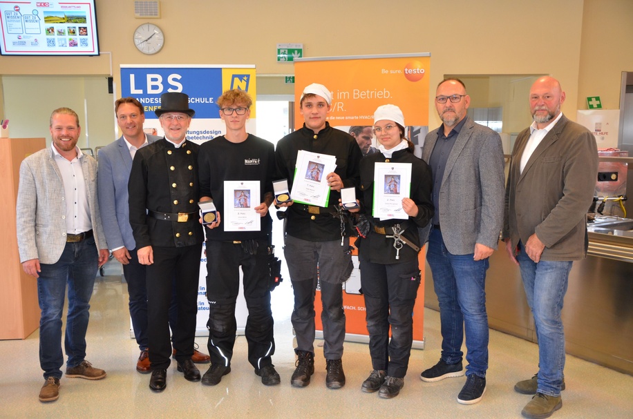 Gruppenfoto einer Preisverleihung mit drei ausgezeichneten Personen, Urkunden und Medaillen.