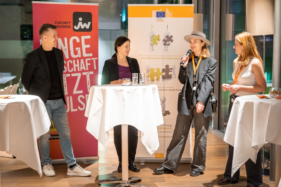 Zu sehen sind Personen, eine davon hat ein Mikrofon in der Hand. Im Hintergrund sind Roll-ups, auf einem steht "Junge Wirtschaft".