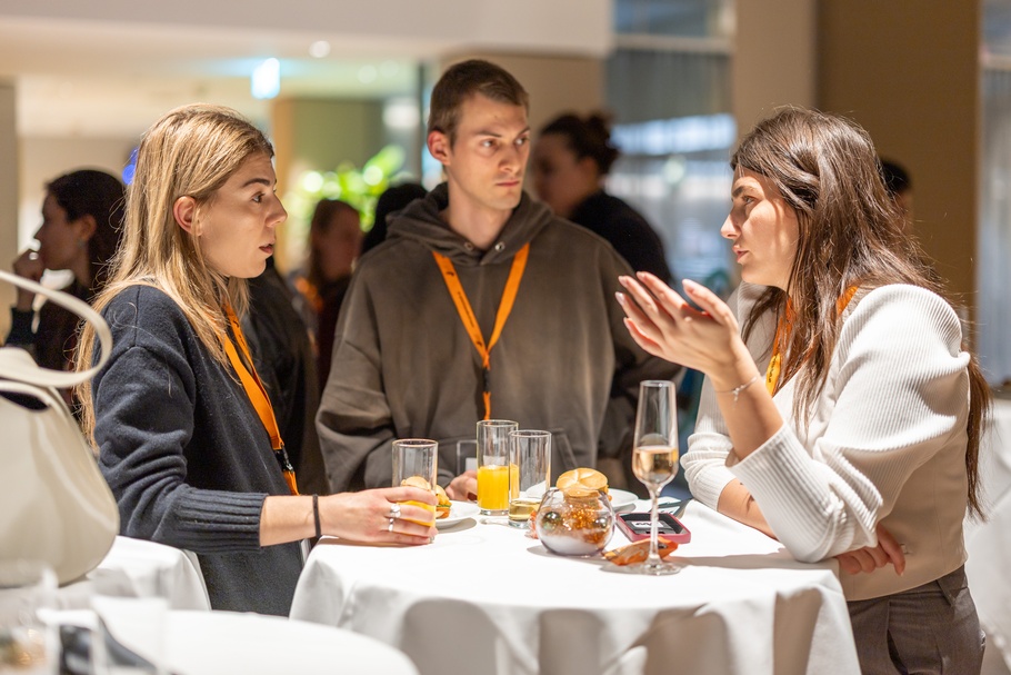 Zu sehen sind Personen, die an einem runden Tisch stehen. Auf diesem sind Gläser und Snacks  platziert. 
