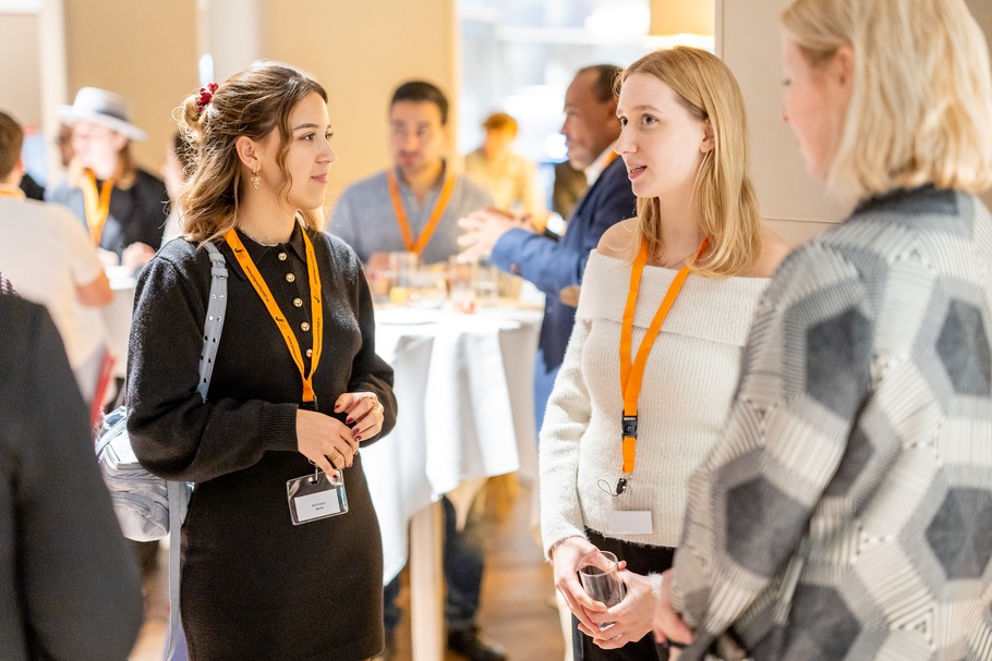 Zu sehen sind Personen, die sich bei einer Veranstaltung unterhalten. Im Hintergrund sind Umrisse anderer Personen und runde Tische zu erkennen. 